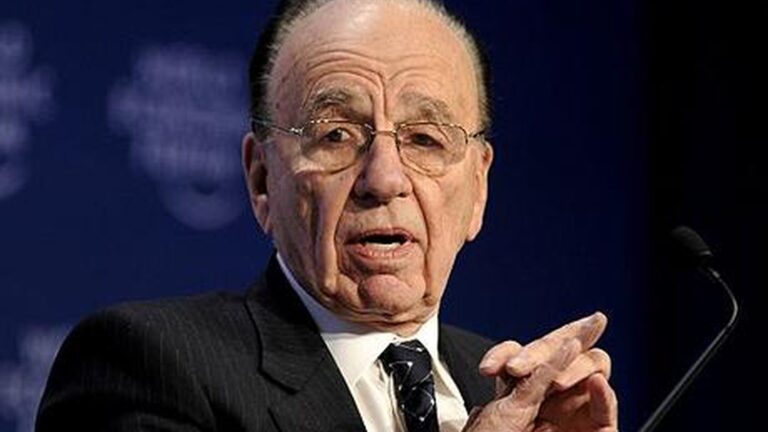 Rupert Murdoch wearing glasses and a suit speaking at a conference with a microphone