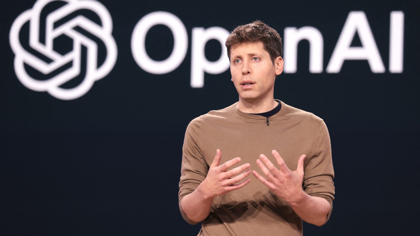 OpenAI CEO Sam Altman speaking on stage in front of the OpenAI logo