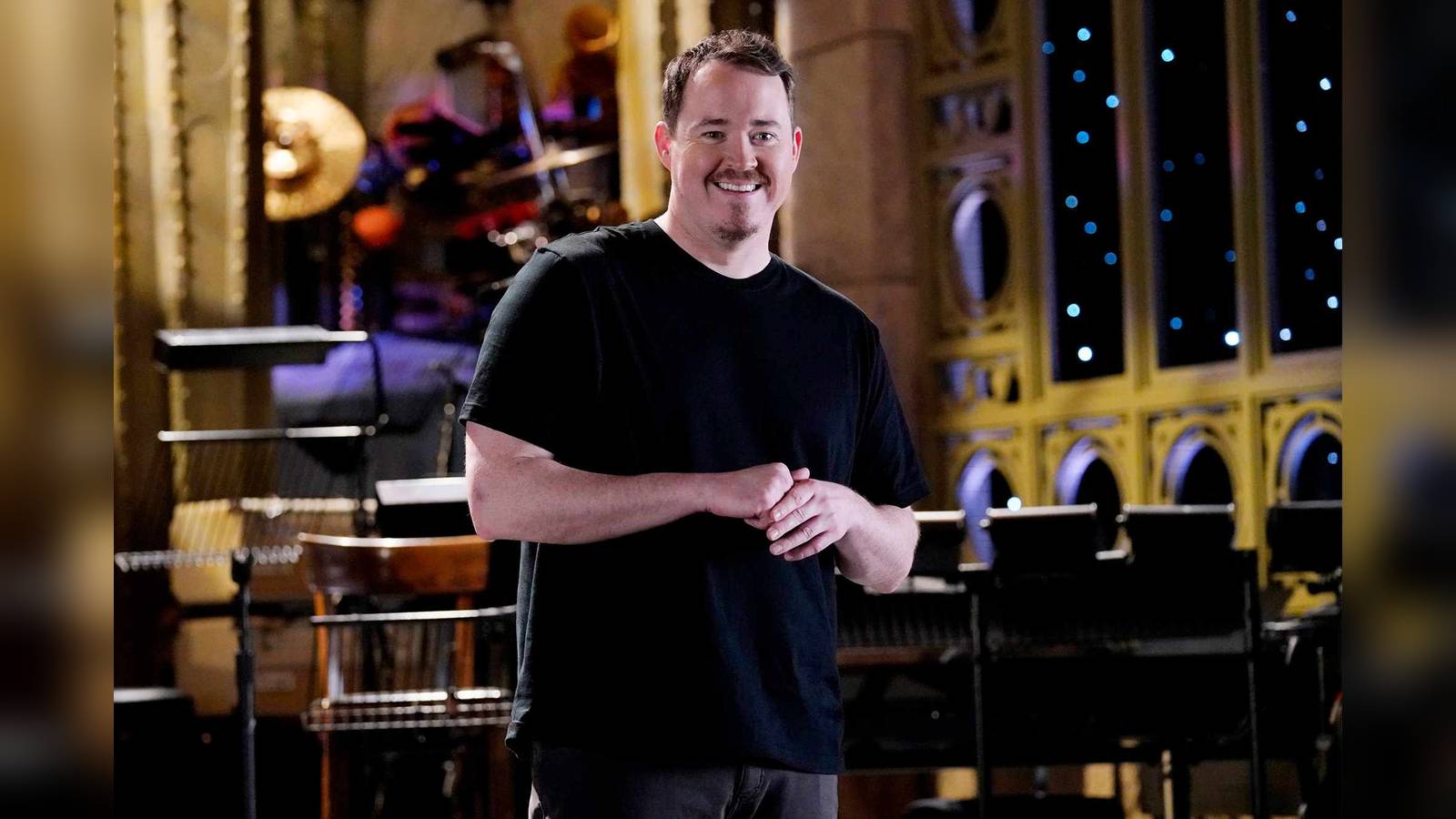Comedian Shane Gillis smiling on the Saturday Night Live stage wearing a black t-shirt
