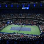 Wide angle view of a large indoor tennis stadium with players on court and packed stands