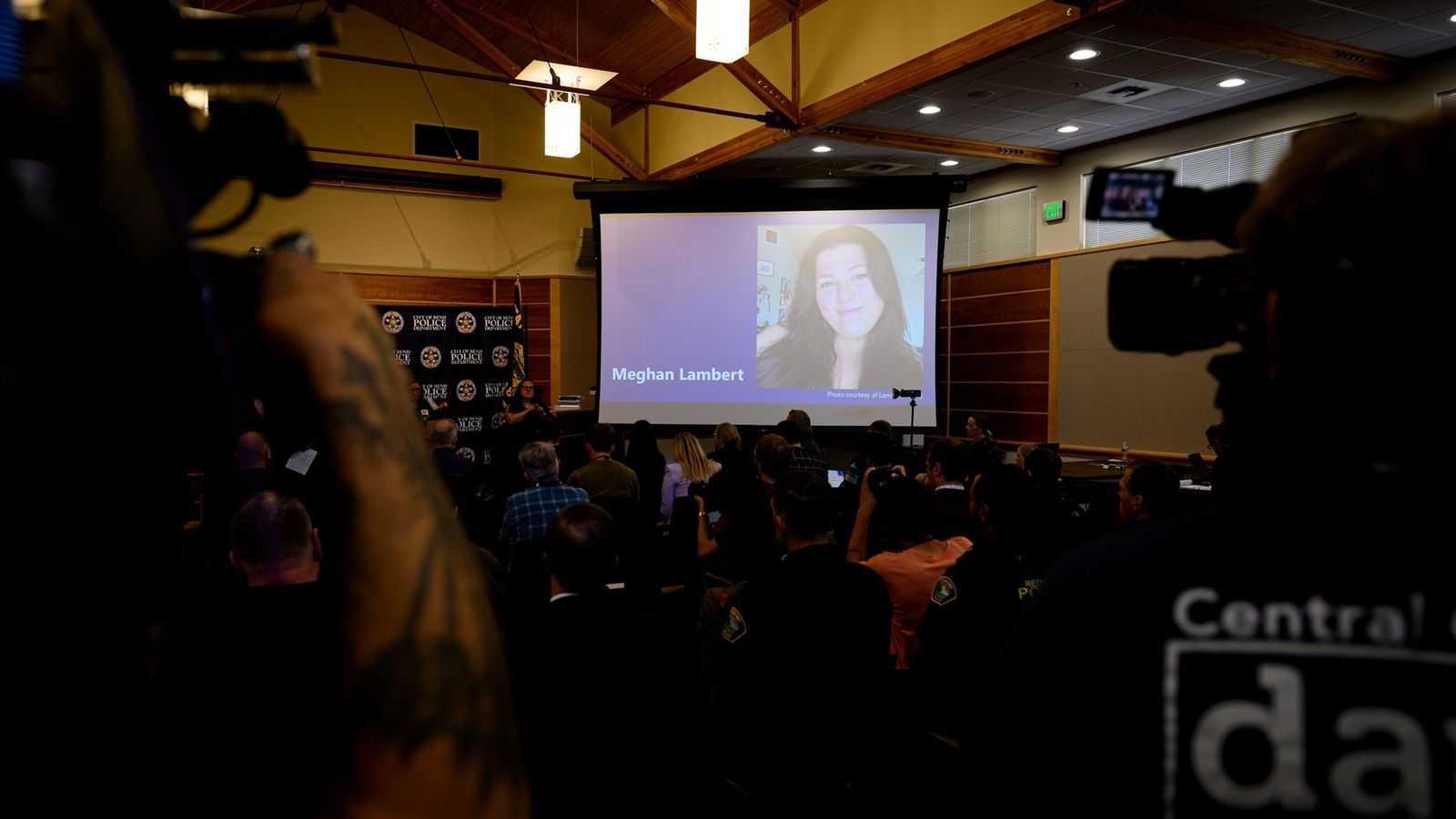 Audience watching a presentation screen with a victim photo at a press conference