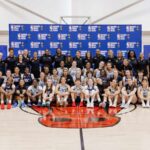 Group of young female basketball players and coaches posing in front of a branded backdrop on a cour