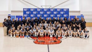 Group of young female basketball players and coaches posing in front of a branded backdrop on a cour