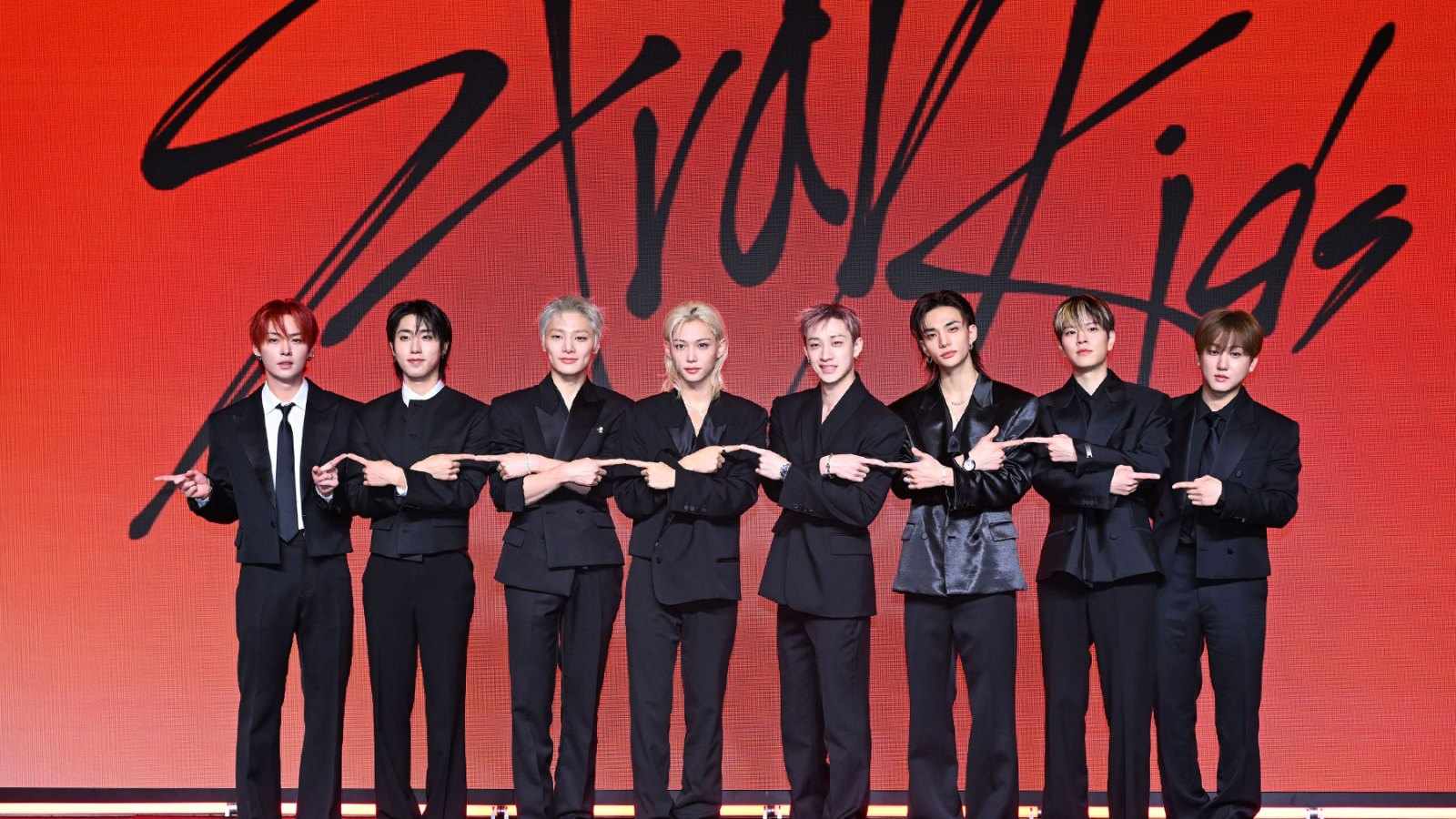 Members of the K-pop group Stray Kids posing together in black suits against a red background