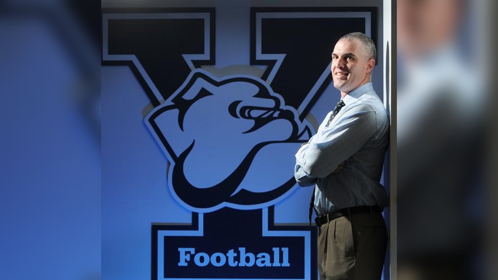 Former Yale football coach Tony Reno posing in front of a Yale Football wall logo