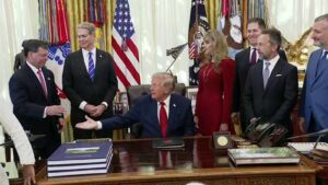 President Trump seated at the Resolute Desk in the Oval Office with officials