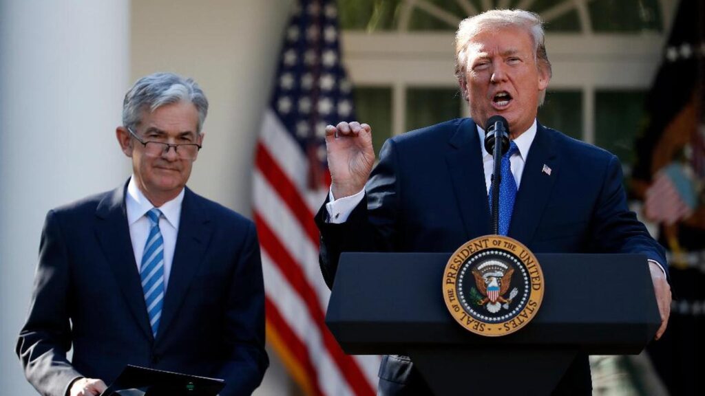 Donald Trump speaking at a podium with Jerome Powell standing nearby in front of American flags