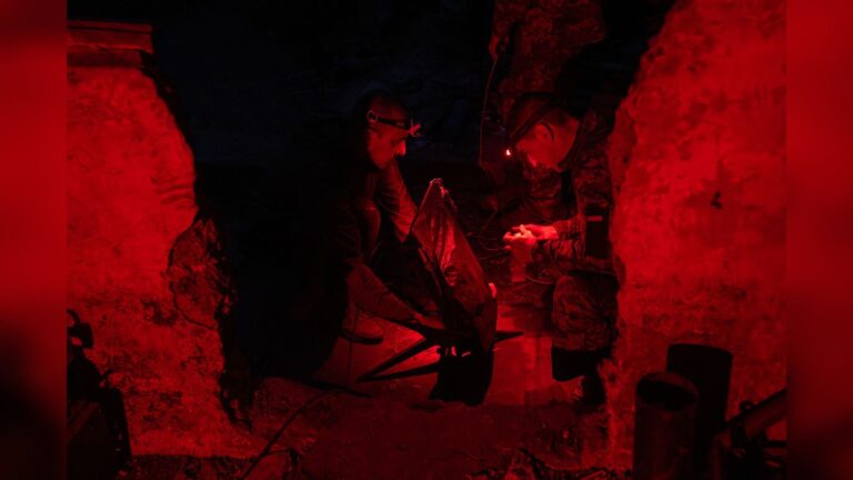 Two Ukrainian soldiers in camouflage gear operating a Starlink satellite terminal under red light